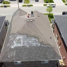 Person cleaning roof with a power washer; left side cleaner than right. Rooftop view of three houses on a suburban street.