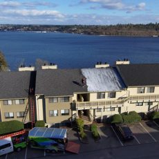 A person is cleaning the roof of a beige apartment building near a large body of water with vehicles parked below.